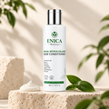 A bottle of ENICA Wellness Argan, Biotin & Collagen Hair Conditioner stands on a textured stone pedestal with a dollop of white conditioner and green leaves in a softly lit, natural setting.